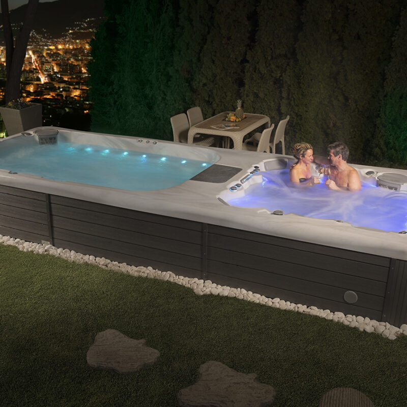 A couple soaks in the spa portion of a luxurious swim spa at night. The spa is beautifully illuminated with blue and purple lights from the LED system which demonstrates the perfect reason you can be thankful for your swim spa.