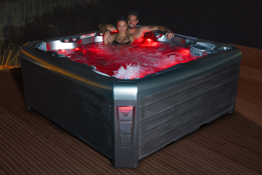 A man and woman sit nestled together in a hot tub lit with red LED lighting to enhance their hot tub date night.