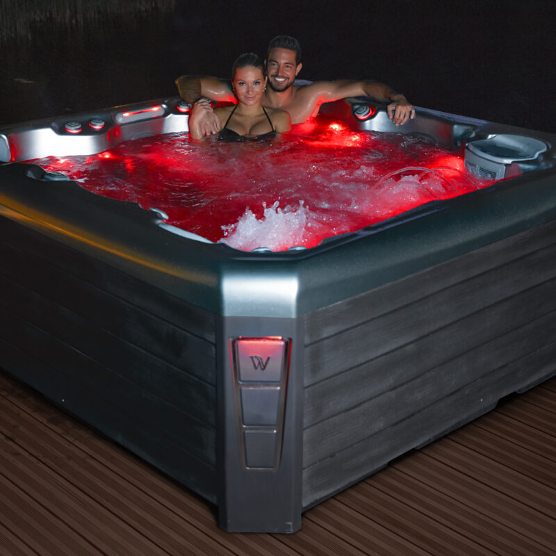 A man and woman sit nestled together in a hot tub lit with red LED lighting to enhance their hot tub date night.