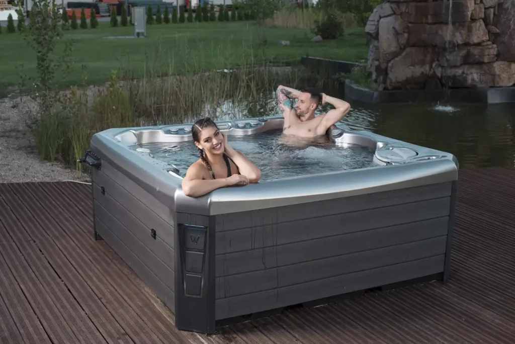 A man and woman relax in a Wellis Hot Tub on a dock next to a pond.